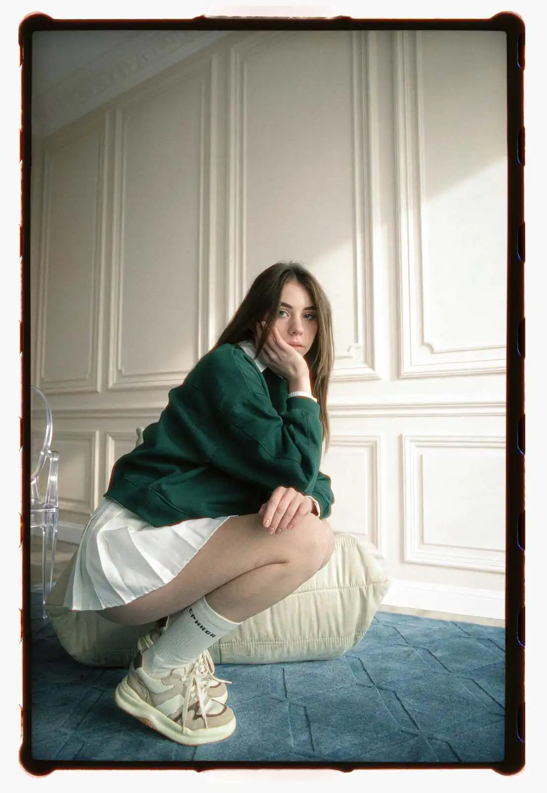 Young woman crouching indoors against classic wall paneling wearing a green sweatshirt white skirt and sneakers photographed on analog film with soft natural window light