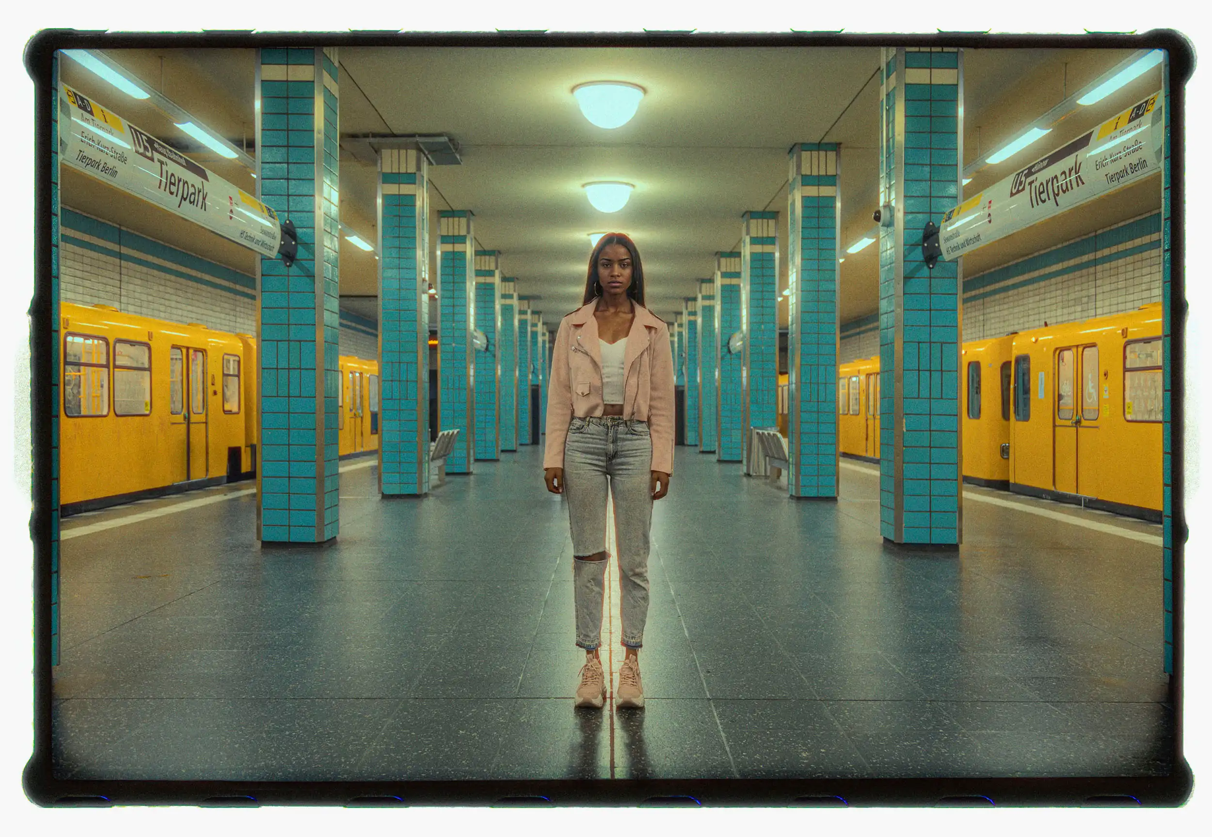 Young woman standing centered on an empty subway platform between yellow trains photographed on analog film with symmetrical tiled columns and soft artificial light