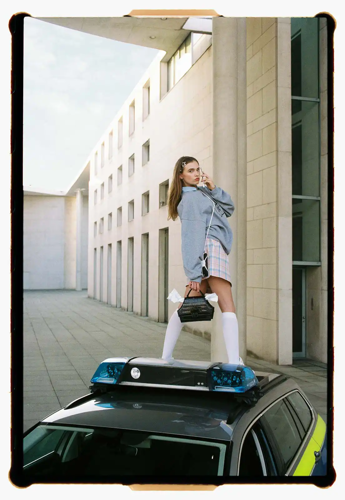 Young woman standing on the roof of a police car in an urban setting wearing a hoodie short skirt and knee high socks holding a handbag and phone photographed on analog film with modern architecture in the background