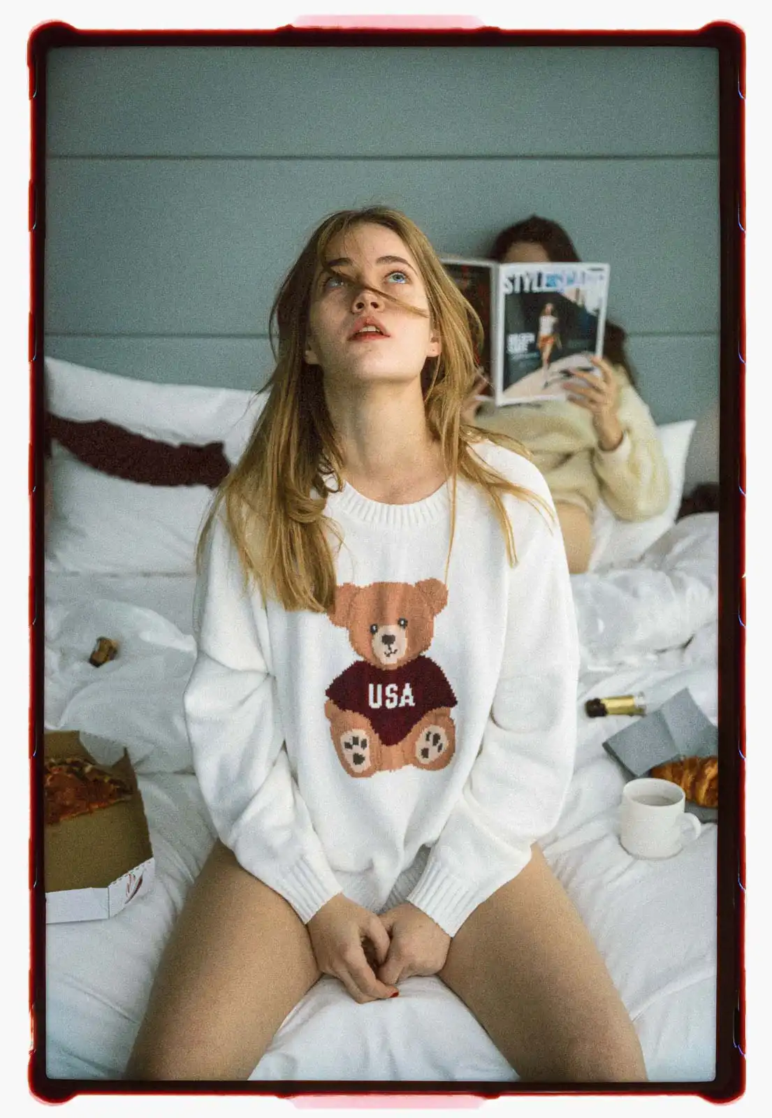 Young woman sitting on an unmade bed wearing an oversized sweater by Hollister Co with a teddy bear motif looking upward with a distant expression photographed on analog film in a softly lit interior scene