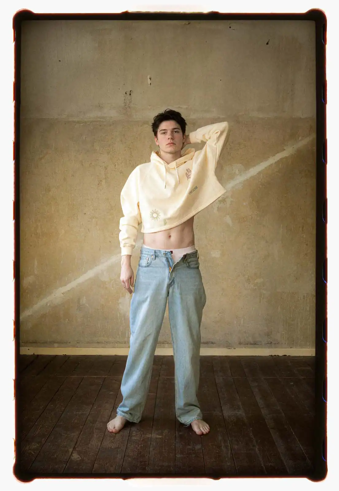 Young person standing barefoot in a worn interior wearing loose denim jeans and a cropped hoodie photographed on analog film with soft natural light and a textured wall behind