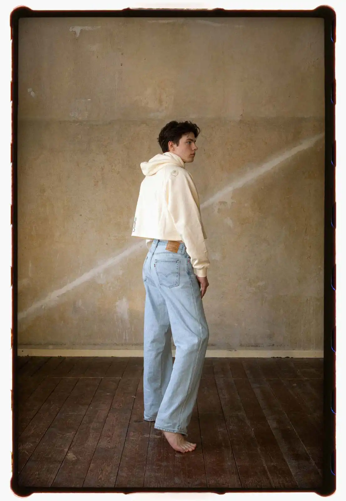 Young man standing barefoot in a minimal interior space wearing light blue jeans and a cropped hoodie photographed on analog film with soft natural light and a textured wall in the background