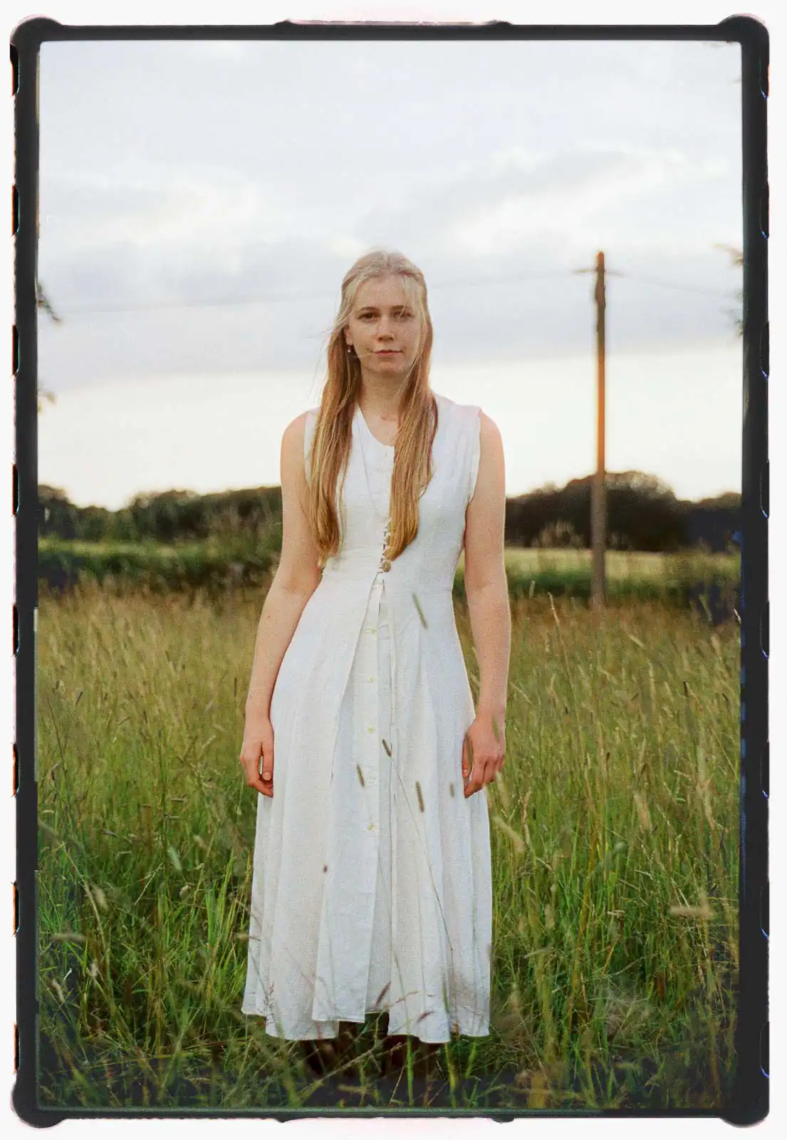 Young woman standing alone in a grassy field at dusk, wearing a white sleeveless dress, photographed on analog film with soft natural light and visible film borders, conveying quiet presence and vulnerability.