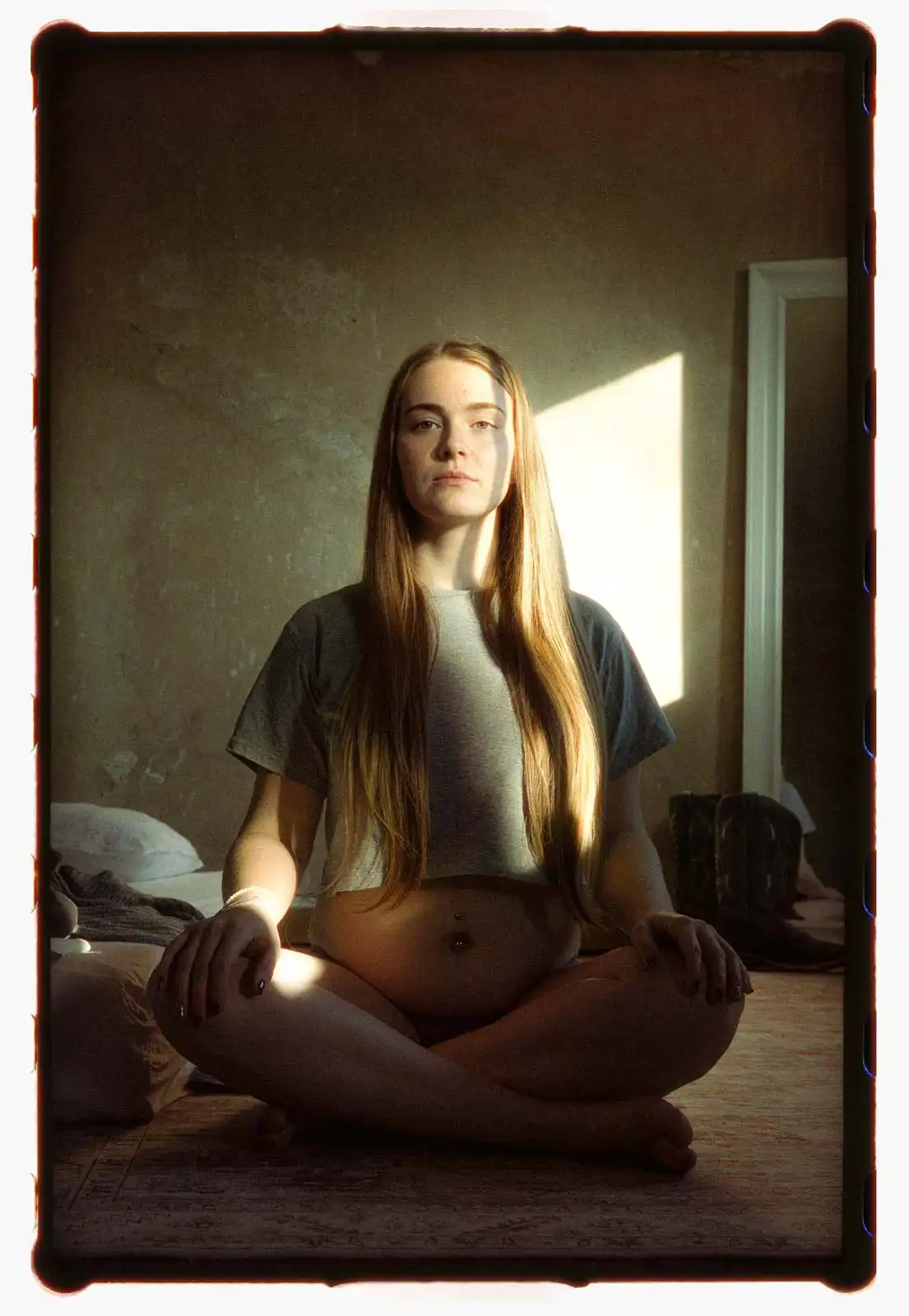 Young pregnant woman standing barefoot in a worn interior room wearing casual clothing photographed on analog film with soft window light and a mirror reflecting the space behind her