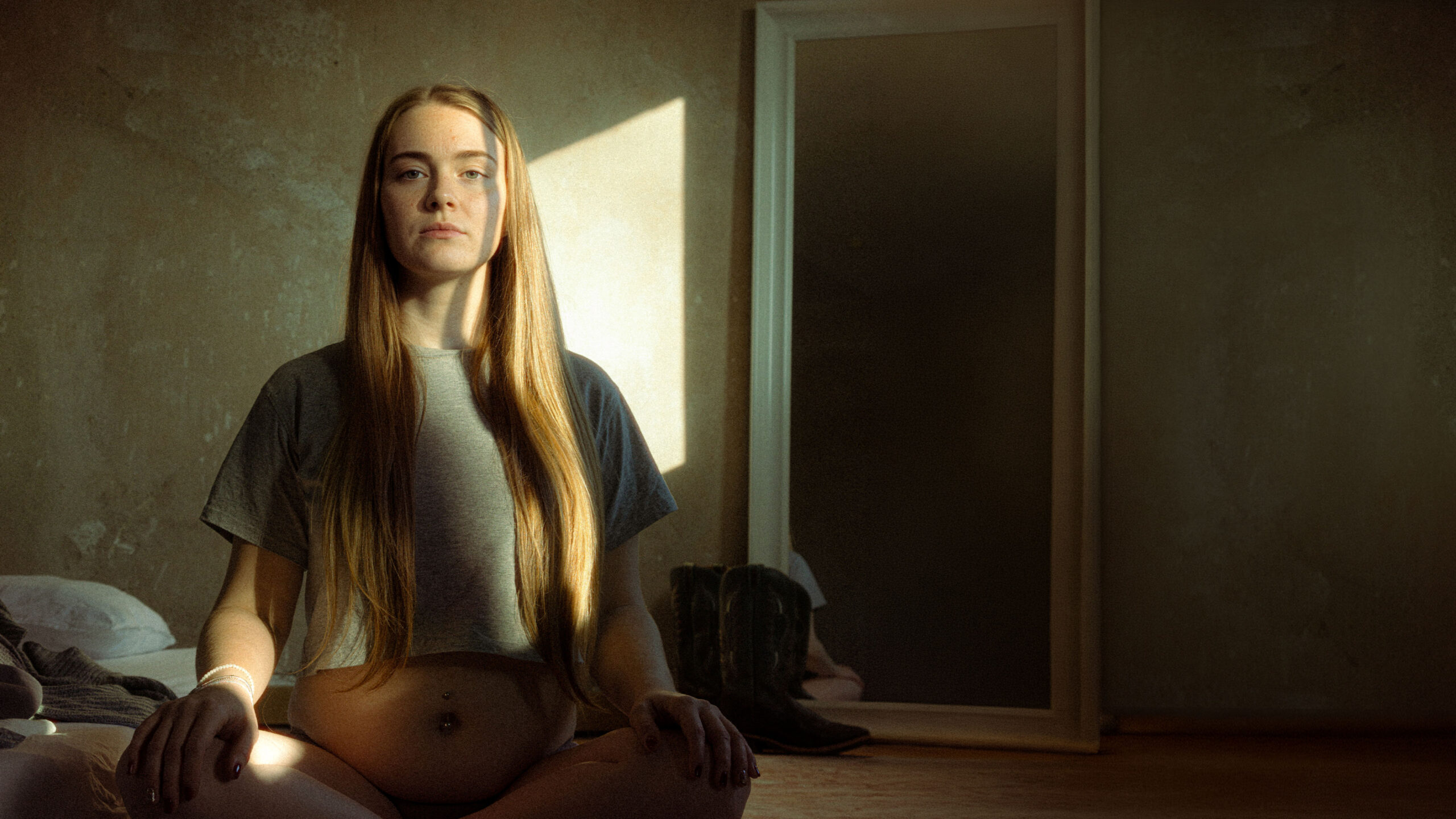 Young pregnant Gen Z woman sitting cross legged on floor in empty apartment shaped by dramatic winter sunlight