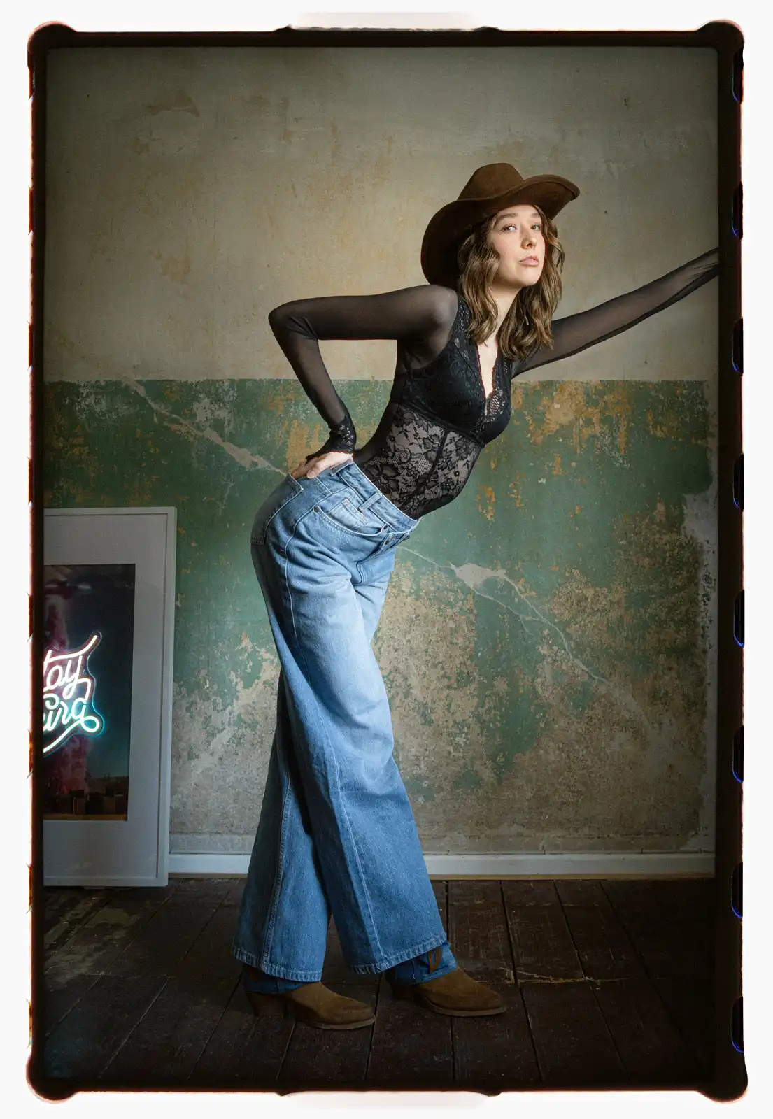 Young woman wearing lace bodysuit wide leg jeans and cowboy hat posing confidently against textured wall portrait photography