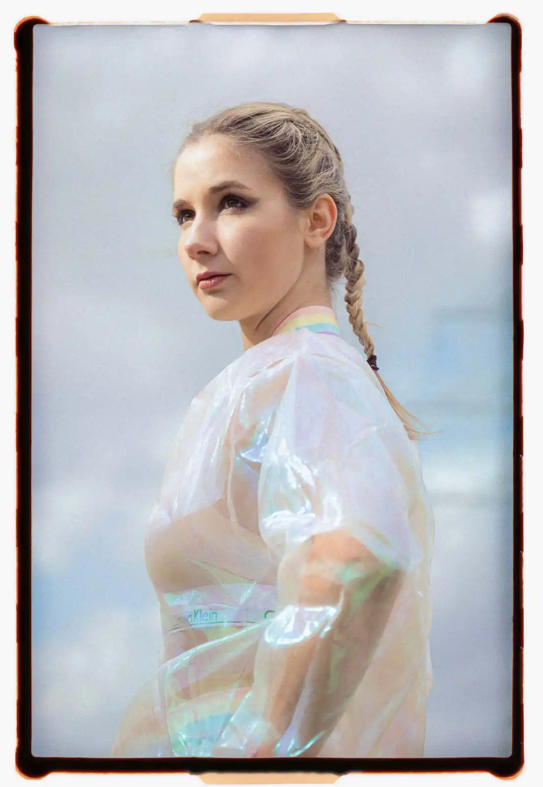 Young woman photographed in close portrait view outdoors wearing a translucent iridescent top with braided hair and soft makeup captured on analog film against a cloudy sky