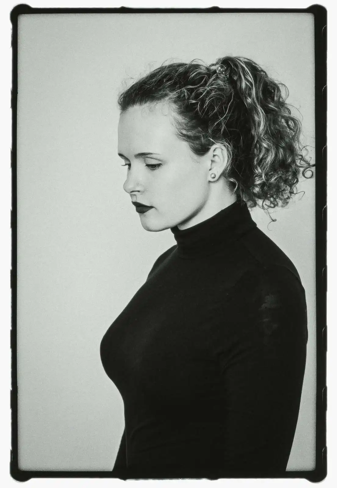 Young woman shown in side profile portrait wearing a dark high neck top with curly hair tied back photographed in black and white on analog film against a plain studio background