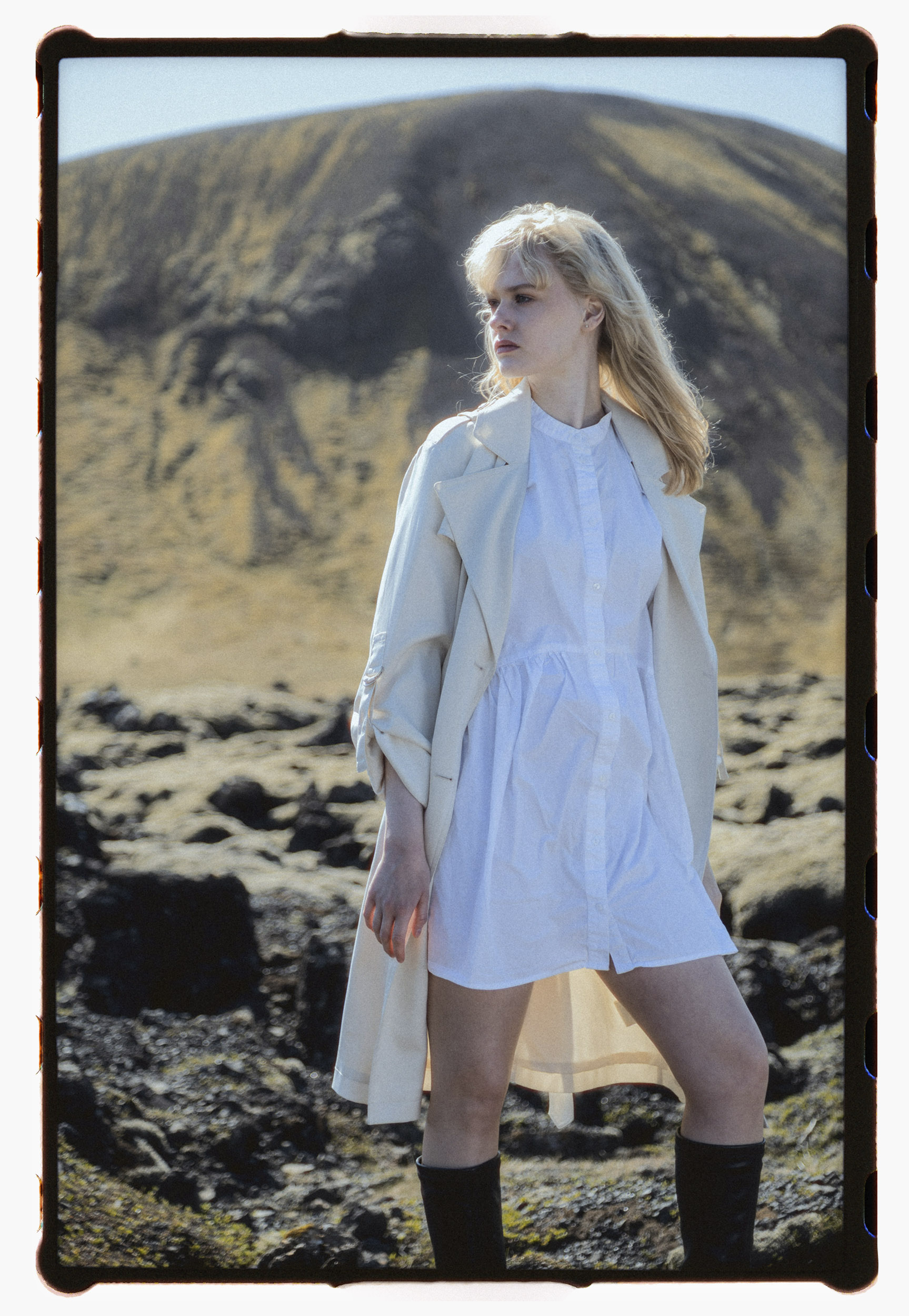 Analog fashion portrait of a young blonde woman in a white shirt dress and light trench coat, standing in front of a dark volcanic mountain in Iceland. She looks to the side as the wind lifts her hair. The rocky lava field beneath her contrasts with the softness of her expression and outfit.