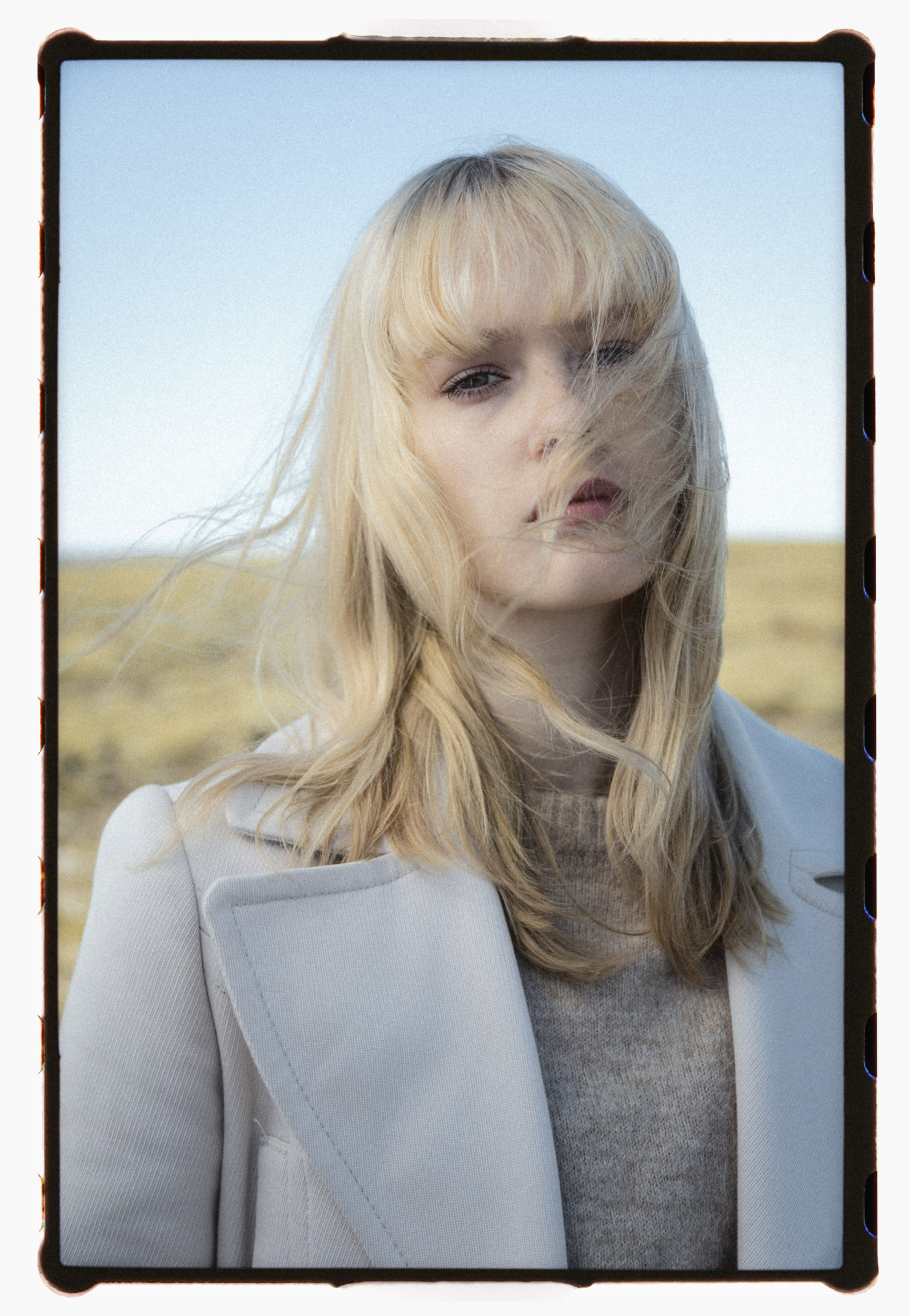 Close-up analog portrait of a blonde woman in a light coat, standing in the Icelandic countryside. Her windswept hair partially veils her face, creating a soft, ethereal atmosphere with a hint of defiance.