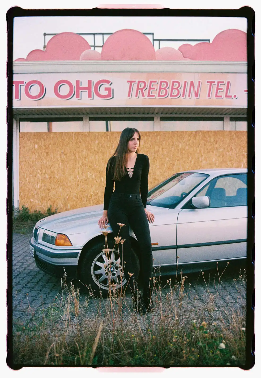 Young woman standing beside a silver car in a semi rural urban edge setting wearing a black fitted outfit photographed on analog film with overgrown grass and a closed storefront in the background