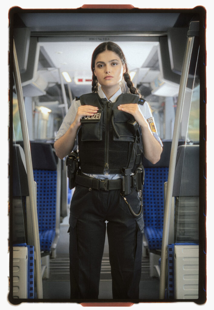 Analog film portrait of a young German policewoman standing confidently in the aisle of a German regional train. She wears a black protective vest over her uniform, with braided hair and a composed, direct gaze. The scene is staged but realistic, evoking authority, youth, and subtle cinematic flair.