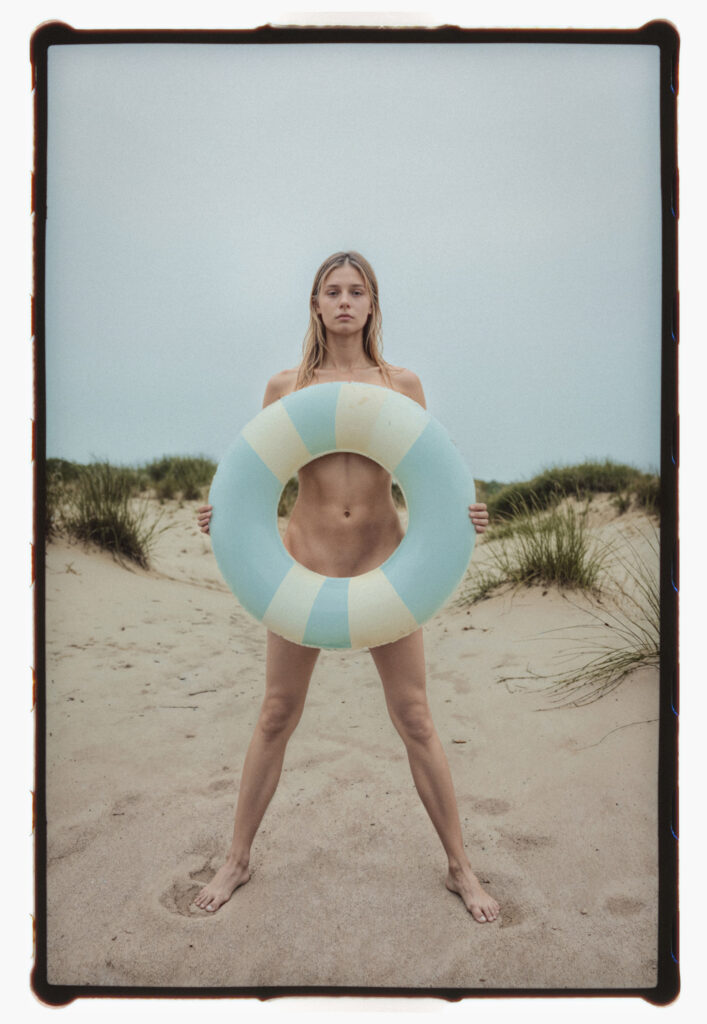 Analog film photo of a blonde naked woman standing confidently on a sandy beach, holding a large pastel-colored swim ring in front of her body. She stands barefoot, legs apart, surrounded by dune grass under a soft blue sky. The image captures youth, summer freedom, and body neutrality with an editorial, artful tone.