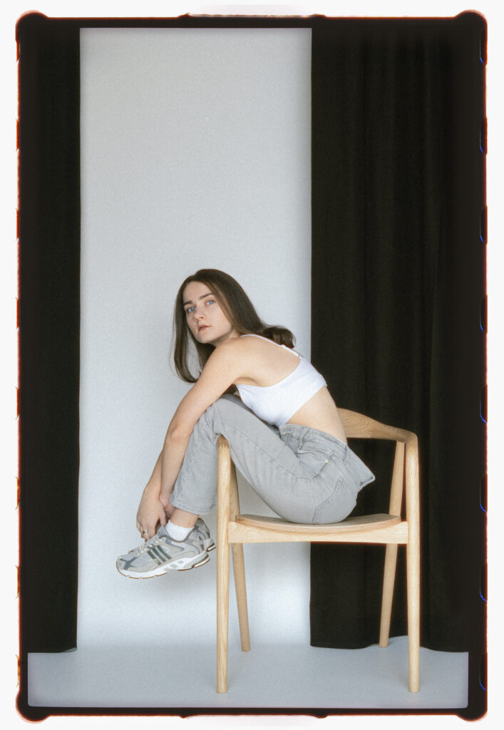 Analog studio portrait of a young woman sitting sideways on a light wooden chair. She wears a cropped white tank top, light grey jeans, and chunky Adidas sneakers. The backdrop is a minimal black-and-white curtain setup, emphasizing clean lines, natural posture, and a quiet moment of self.
