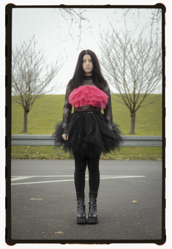 Analog portrait titled Ornament of a woman in a black lace outfit with a bold pink ruffle, standing on a road with bare trees and a grassy hill in the background.