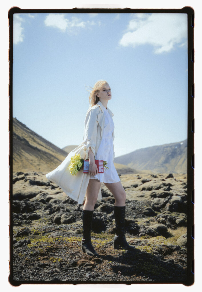 Analog portrait of a young woman standing on a volcanic landscape in Iceland, holding a bouquet of yellow flowers and two red-and-white cans of Monster Energy drink. She wears a white dress, a light trench coat, and black leather boots. Her hair blows in the wind beneath a bright blue sky, mountains fading in the distance. Shot on film.