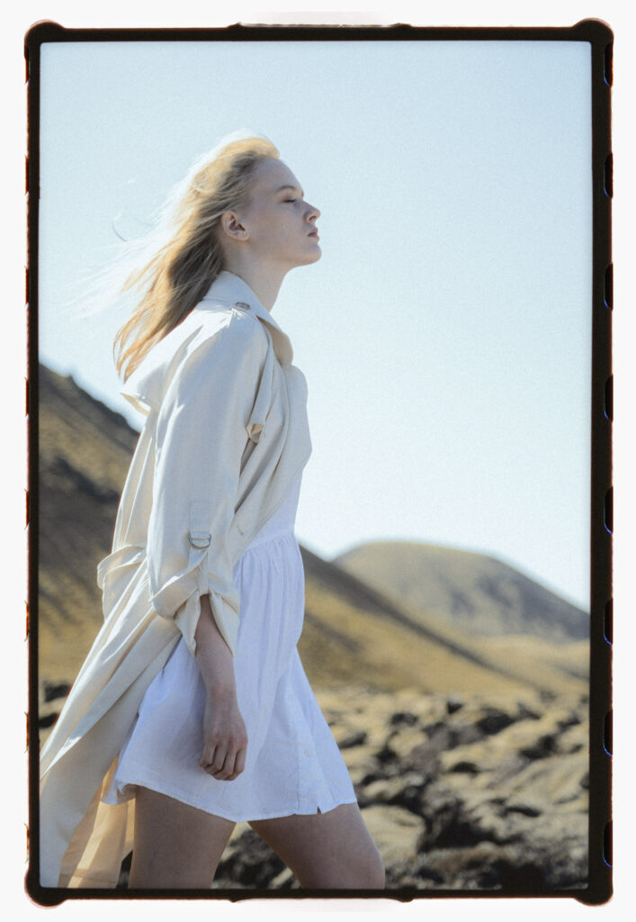 Side-profile analog portrait of a blonde woman in a flowing white dress and light trench coat, walking through an Icelandic lava field with rugged hills in the background. Her hair is swept back by the wind as she gazes ahead with calm determination.