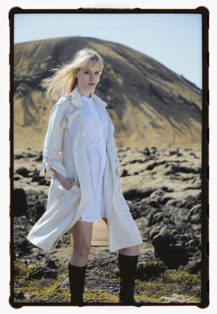 Blonde woman in a light trench coat and white dress, standing in front of a volcanic hill in Iceland. Her stance is grounded and powerful, with hands in her pockets and wind-blown hair.